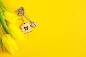 Key ring in the shape of wooden house with key on yellow background and spring tulips. Building, design, project, moving to new home, mortgage, rent and purchase real estate, summer offer. Copy space photo