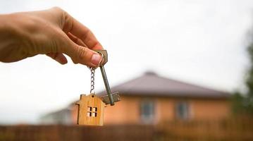 Hand with a key and a wooden key ring-house. Background of fence and cottage. Building, project, moving to a new home, mortgage, rent and purchase real estate. To open the door. Copy space photo