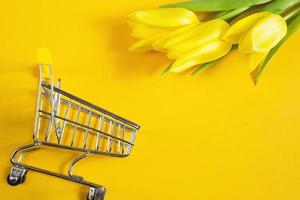 tulipanes amarillos sobre un fondo de color sólido y carrito de compras. compras en línea en primavera y verano, compra de regalos para las vacaciones, semana santa, día de la mujer, 8 de marzo. floristería, floristería. copie el espacio foto