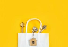 Key ring in the shape of wooden house with key on a yellow background in a white paper gift wrapping bag. Mortgage, rent and purchase real estate, beneficial offer for construction. Copy space photo