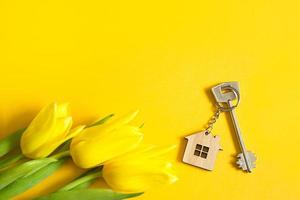 Key ring in the shape of wooden house with key on yellow background and spring tulips. Building, design, project, moving to new home, mortgage, rent and purchase real estate, summer offer. Copy space photo