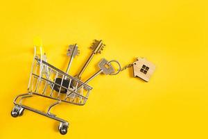 Key ring in the shape of wooden house with key on a yellow background and shopping cart. Mortgage, rent and purchase real estate, spring and summer offer for construction. Copy space photo