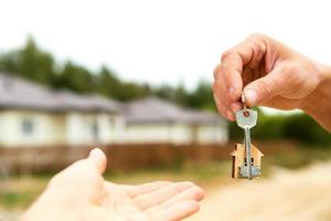 Hand with a key and a wooden key ring-house. Background of fence and cottage. Building, project, moving to a new home, mortgage, rent and purchase real estate. To open the door. Copy space photo