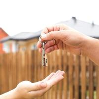Hand with a key and a wooden key ring-house. Background of fence and cottage. Building, project, moving to a new home, mortgage, rent and purchase real estate. To open the door. Copy space photo