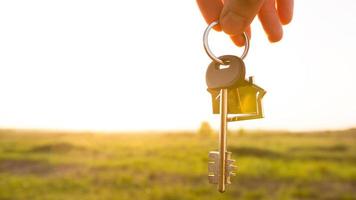 House key with keychain in hand. Background of sky, sunlight and field. Dream of home, building a cottage in countryside, plan and project design, farm, moving to a new home, accommodation. Copy space photo