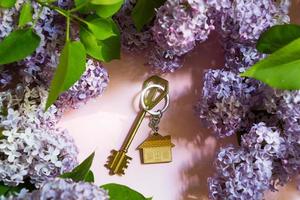 House key with keychain on pink spring background and lilac branches. Summer holiday home, booking cottage in the countryside, moving to new home, mortgage, rent and purchase real estate. Copy space photo