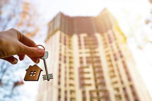 Hand with a key and a wooden key ring-house. Background of multi-apartment skyscraper. Building, moving to a new flat, mortgage, rent and purchase real estate. To open the door. Copy space photo