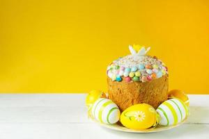 Easter cake with painted eggs on a platter in a white table. Traditional festive food. Yellow background photo