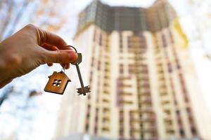 Hand with a key and a wooden key ring-house. Background of multi-apartment skyscraper. Building, moving to a new flat, mortgage, rent and purchase real estate. To open the door. Copy space photo