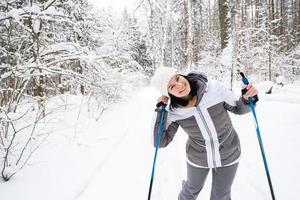 Skier a woman in a membrane jacket with ski poles in his hands with his back against the background of a snowy forest. Cross-country skiing in winter forest, outdoor sports, healthy lifestyle. photo
