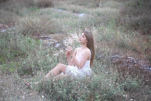 hermosa mujer joven sentada en el campo de hierba verde y soplando diente de león. al aire libre. disfruta de la naturaleza. niña sonriente saludable en el césped de primavera. concepto libre de alergias. libertad foto