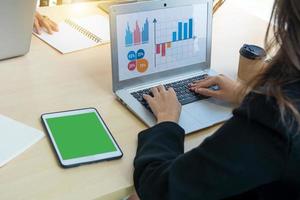 vista posterior joven mujer de negocios sentada en la oficina en el escritorio y usando un teléfono inteligente en el escritorio como computadora portátil y tableta en gráficos y gráficos de pantalla. foto