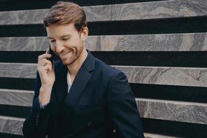 Young attractive confident businessman with neat beard in formal wear speaking on mobile outdoors photo