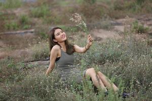 hermosa mujer joven sentada en el campo de hierba verde y soplando diente de león. al aire libre. disfruta de la naturaleza. niña sonriente saludable en el césped de primavera. concepto libre de alergias. libertad foto