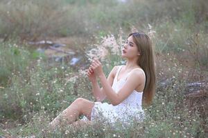 hermosa mujer joven sentada en el campo de hierba verde y soplando diente de león. al aire libre. disfruta de la naturaleza. niña sonriente saludable en el césped de primavera. concepto libre de alergias. libertad foto