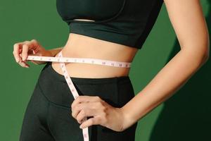Crop close up body of sporty woman measuring her thin waist with a tape measure isolated on green background. Dynamic movement. Strength and motivation. photo