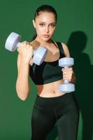 Beautiful strong cheerful young Asian sports woman does exercises with dumbbell isolated on green background. Fitness girl in sportswear posing indoors. Movement, Strength and motivation.