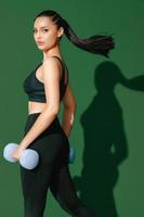 Beautiful strong cheerful young Asian sports woman does exercises with dumbbell isolated on green background. Fitness girl in sportswear posing indoors. Movement, Strength and motivation. photo