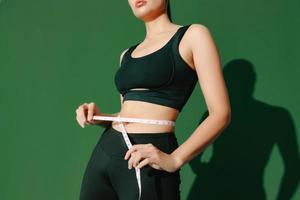 Crop close up body of sporty woman measuring her thin waist with a tape measure isolated on green background. Dynamic movement. Strength and motivation. photo
