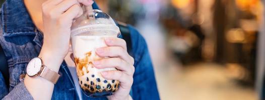 Young girl in denim jacket is drinking brown sugar flavored tapioca pearl bubble milk tea with glass straw in night market of Taiwan, close up, bokeh photo