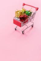 Annual sale, Christmas shopping season concept - mini red shop cart trolley full of gift box isolated on pale pink background, copy space, close up photo