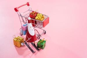 Christmas shopping concept, mini red shop cart trolley with Santa Claus toy and gift box isolated on pale pink background, blank copy space, close up photo