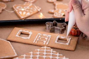 la mujer está decorando la casa de galletas de jengibre con cobertura de crema de hielo blanco sobre fondo de mesa de madera, papel para hornear en la cocina, primer plano, macro. foto