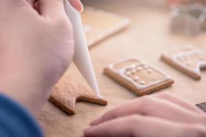 la mujer está decorando la casa de galletas de jengibre con cobertura de crema de hielo blanco sobre fondo de mesa de madera, papel para hornear en la cocina, primer plano, macro. foto