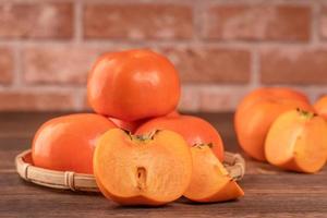 Sliced sweet persimmon kaki in a bamboo sieve basket on dark wooden table with red brick wall background, Chinese lunar new year fruit design concept, close up. photo