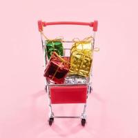 venta anual, concepto de temporada de compras navideñas - mini carrito de compras rojo lleno de caja de regalo aislado en fondo rosa pálido, espacio de copia, primer plano foto