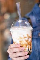 mujer joven está sosteniendo, bebiendo té de leche con burbujas de perlas de tapioca con sabor a azúcar moreno con paja de vidrio en el mercado nocturno de taiwán, de cerca, bokeh foto