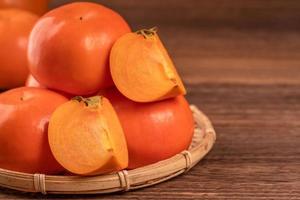 Sliced sweet persimmon kaki in a bamboo sieve basket on dark wooden table with red brick wall background, Chinese lunar new year fruit design concept, close up. photo