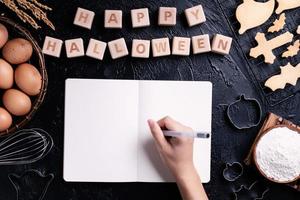 mujer joven está leyendo y escribiendo recetas para hacer galletas de halloween, concepto de diseño de preparación para la fiesta de halloween, vista superior, puesta plana, arriba. foto