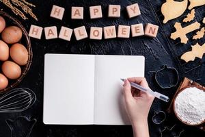 Young woman is reading and writing recipe of making Halloween cookies, design concept of preparing for Halloween party, top view, flat lay, overhead. photo