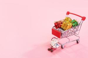 Christmas shopping concept, mini red shop cart trolley with Santa Claus toy and gift box isolated on pale pink background, blank copy space, close up photo