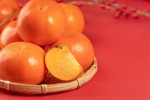 Fresh beautiful sliced sweet persimmon kaki isolated on red table background and bamboo sieve, Chinese lunar new year design concept, close up. photo