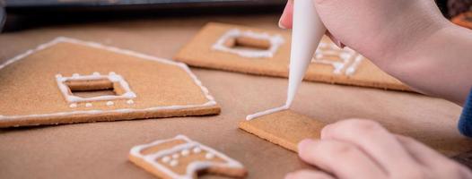 Woman is decorating gingerbread cookies house with white frosting icing cream topping on wooden table background, baking paper in kitchen, close up, macro. photo