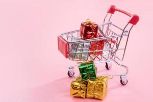 venta anual, concepto de temporada de compras navideñas - mini carrito de compras rojo lleno de caja de regalo aislado en fondo rosa pálido, espacio de copia, primer plano foto