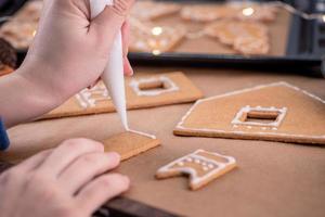 la mujer está decorando la casa de galletas de jengibre con cobertura de crema de hielo blanco sobre fondo de mesa de madera, papel para hornear en la cocina, primer plano, macro. foto