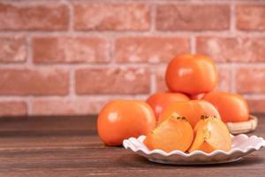 Sliced sweet persimmon kaki in a bamboo sieve basket on dark wooden table with red brick wall background, Chinese lunar new year fruit design concept, close up. photo