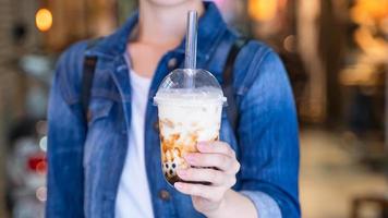 mujer joven está sosteniendo, bebiendo té de leche con burbujas de perlas de tapioca con sabor a azúcar moreno con paja de vidrio en el mercado nocturno de taiwán, de cerca, bokeh foto