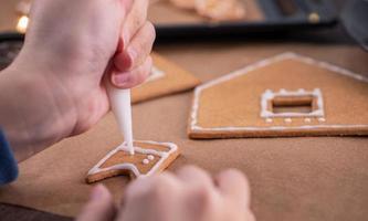 Woman is decorating gingerbread cookies house with white frosting icing cream topping on wooden table background, baking paper in kitchen, close up, macro. photo