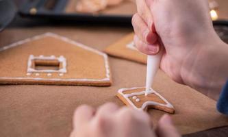 la mujer está decorando la casa de galletas de jengibre con cobertura de crema de hielo blanco sobre fondo de mesa de madera, papel para hornear en la cocina, primer plano, macro. foto