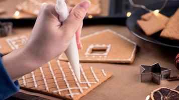 la mujer está decorando la casa de galletas de jengibre con cobertura de crema de hielo blanco sobre fondo de mesa de madera, papel para hornear en la cocina, primer plano, macro. foto