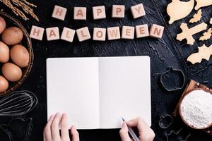 Young woman is reading and writing recipe of making Halloween cookies, design concept of preparing for Halloween party, top view, flat lay, overhead. photo