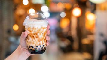 Young woman is holding, drinking brown sugar flavored tapioca pearl bubble milk tea with glass straw in night market of Taiwan, close up, bokeh photo