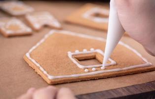 la mujer está decorando la casa de galletas de jengibre con cobertura de crema de hielo blanco sobre fondo de mesa de madera, papel para hornear en la cocina, primer plano, macro. foto