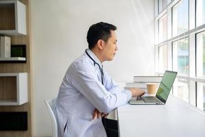 médico masculino asiático usando una computadora portátil junto a la ventana en su oficina. foto