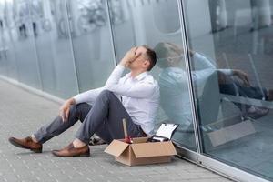 The fired office worker fell to his knees and covered his face due to stress. In front of him is a cardboard box with stationery. The man is unhappy due to a reduction in the robot. photo