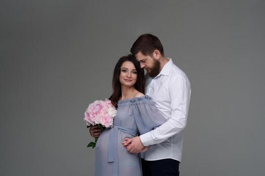A pregnant woman and her husband are hugging on a gray background in the studio. photo